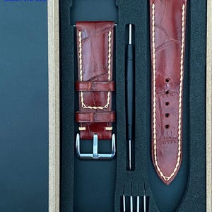 Può includere: Un set di cinturini per orologi in una scatola. Il set include due cinturini per orologi in pelle marrone rossiccio con cuciture bianche, un attrezzo nero e diversi piccoli perni metallici. I cinturini hanno un motivo strutturato. La scatola ha il testo "Leather Art Shop".