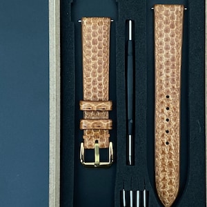 Op de afbeelding: Een horlogebandset in een presentatiedoos. De band is lichtbruin met een textuurpatroon, een goudkleurige gesp en een zwart gereedschap. De doos bevat ook kleine metalen pinnen. De tekst "Leather Art Shop" is zichtbaar.