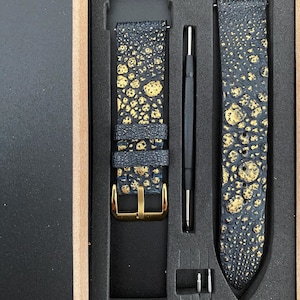Puede incluir: Un juego de correas de reloj en una caja de madera. Las correas son negras con patrones irregulares de color dorado. Se ve una hebilla dorada. También se incluyen una herramienta negra y dos pasadores plateados. El texto "Leather Art Shop" es visible.