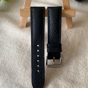 Pode incluir: Duas pulseiras de relógio de couro preto com superfície texturizada. Uma pulseira tem uma fivela prateada e a outra tem vários orifícios de ajuste. As pulseiras são exibidas em um pequeno cavalete de madeira.
