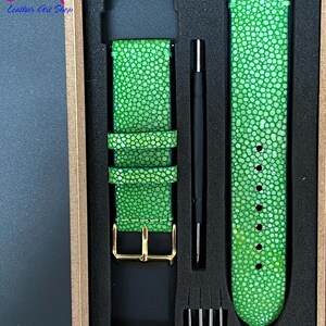 May include: A vibrant green watch strap with a textured finish, displayed in a box. The strap features a gold-coloured buckle and comes with a tool for fitting. The box also contains spare pins.