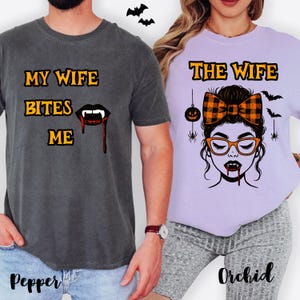 Puede incluir: Dos camisetas con temática de Halloween. Una es gris oscuro con el texto "MY WIFE BITES ME" y un gráfico de boca de vampiro. La otra es de color lila claro con "THE WIFE" y un gráfico de mujer vampiro. También aparecen murciélagos y una calabaza.