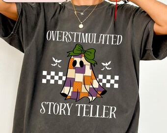 Op de afbeelding: Donkergrijs t-shirt met de witte woorden "OVERSTIMULATED STORY TELLER". Het shirt heeft een cartoon spook met een patchwork ontwerp in oranje, paars en bruin, een groene strik en twee kleine vleermuizen. Een wit en zwart geblokt patroon staat eronder.