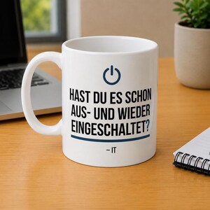 Könnte beinhalten: Ein weißer Keramikbecher mit einem dunkelblauen Power-Button-Symbol oben, gefolgt von dem schwarzen Text "HAST DU ES SCHON AUS- UND WIEDER EINGESCHALTET?" mit einer dunkelblauen horizontalen Linie darunter und dem Text "- IT" am unteren Rand.