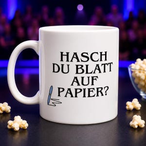 Könnte beinhalten: Ein weißer Keramikbecher mit dem schwarzen Aufdruck "HASCH DU BLATT AUF PAPIER?" und einer kleinen Grafik eines blauen Stifts, der eine Linie zeichnet. Der Becher verfügt über einen Standardhenkel und eine glatte, glänzende Oberfläche.