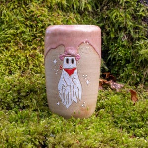 Peut inclure: Une tasse en céramique avec un glaçage rose et un motif peint à la main d'un fantôme portant un chapeau de cowboy et un bandana. Le fantôme est blanc avec des yeux noirs et un bandana rouge.