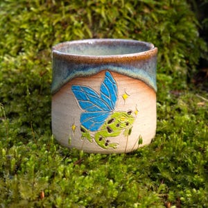 Könnte beinhalten: Eine Keramiktasse mit blauer und weißer Glasur. Die Tasse zeigt ein handgemaltes Design eines grünen Frosches mit blauen Flügeln und Glitzer.