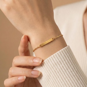 May include: A gold-colored bracelet with a rectangular bar pendant. The bracelet is made of a delicate chain and is worn on a wrist. The background is a neutral beige color.