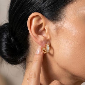 Peut inclure: Boucles d'oreilles créoles dorées avec un insert en émail blanc. Les boucles d'oreilles sont petites, circulaires et portées à l'oreille. L'image montre un gros plan des boucles d'oreilles, soulignant leur design et le contraste entre l'or et le blanc.