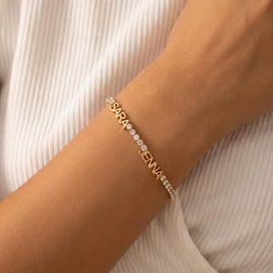 Puede incluir: Pulsera dorada con los nombres "SARA" y "JENNA" escritos en mayúsculas. La pulsera presenta una línea de pequeñas piedras preciosas transparentes. La pulsera se lleva en un brazo.