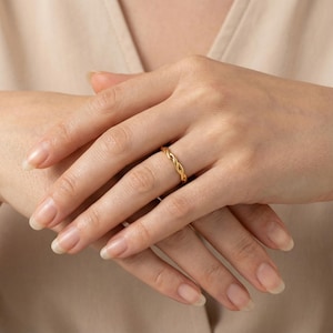 Op de afbeelding: Een goudkleurige ring met een gevlochten ontwerp. De ring wordt aan een vinger gedragen, waarbij het ingewikkelde geweven patroon wordt getoond. De warme tint van de ring contrasteert met de huidskleur, wat de elegante eenvoud benadrukt. Een close-up benadrukt de details van de ring.