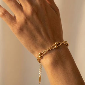 May include: A gold-colored bracelet with a twisted rope design and small ball accents. The bracelet is on a wrist, and the clasp is visible. The jewelry is likely made of stainless steel or gold-plated material.