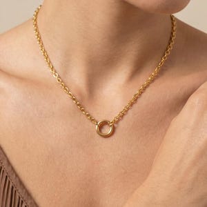 May include: A gold-colored necklace featuring a delicate chain and a circular pendant. The necklace is worn around the neck, showcasing its simple yet elegant design. The chain links are visible, adding texture to the piece.