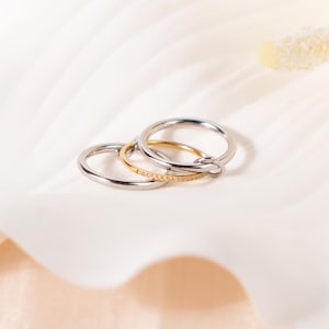 Puede incluir: Tres anillos, dos plateados y uno dorado, están colocados sobre una superficie blanca. Un anillo plateado es liso, el otro tiene un pequeño lazo. El anillo dorado está adornado con pequeñas piedras brillantes. Probablemente sean joyas.