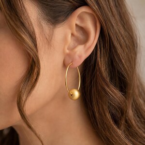 May include: A gold hoop earring featuring a single, smooth, matte-finish gold sphere bead threaded onto the thin circular wire. The earring has a warm, metallic yellow tone and a simple, clean design that hangs vertically from the earlobe.