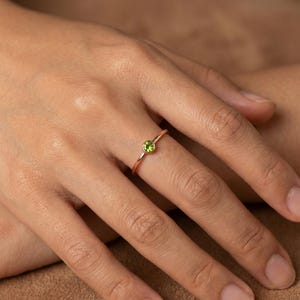 May include: A delicate rose gold ring featuring a round, light green gemstone. The ring has a thin band and a simple, elegant design. The gemstone is set in a bezel setting, and the ring is shown on a person's finger.