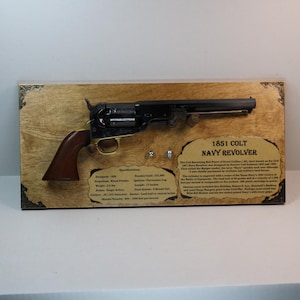 May include: An 1851 Colt Navy revolver mounted on a wooden plaque. The black and gold firearm has a brown wooden grip. The plaque includes text describing the revolver's specifications and history. The plaque is 38 cm long.