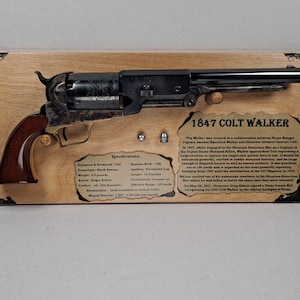 Puede incluir: Una exhibición con un revólver Colt Walker de 1847 montado sobre una placa de madera clara. Incluye dos pequeños proyectiles de metal y texto impreso que detalla la historia y las especificaciones del arma, incluyendo un peso de 2,04 kilogramos.