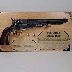 Pode incluir: Revólver Colt Army Modelo 1860 montado numa placa de madeira. A arma de fogo preta e dourada tem uma empunhadura de madeira escura e é acompanhada por duas balas. A placa inclui texto detalhando especificações e informações históricas.