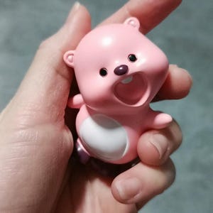 May include: A pink and white figurine of a cartoon animal with a wide open mouth and a single tooth showing. The figurine has black eyes and a purple nose.