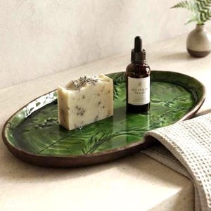 May include: An oval green ceramic tray with a leaf pattern, a rectangular bar of off-white soap with visible dried herbs, and a small brown glass dropper bottle with a white label reading Botanical Elixir, arranged on a light surface.