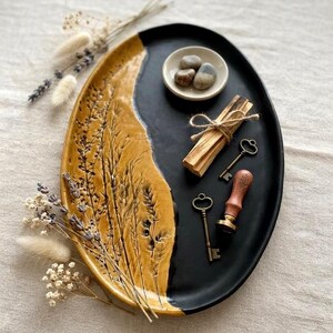 Puede incluir: Una bandeja ovalada de cerámica con diseño dividido, con un lado amarillo mostaza brillante con espigas de trigo en relieve y un lado negro mate. Contiene un cuenco blanco pequeño con tres piedras lisas, un atado de madera, dos llaves de metal y un sello.