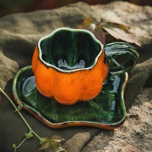 Pode incluir: Um conjunto de chávena e pires em cerâmica com formato de abóbora. A chávena tem um exterior laranja vivo com um interior verde escuro brilhante e uma asa a condizer. O pires tem o formato de uma folha verde com acabamento vidrado craquelado.