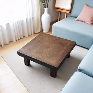 May include: A square, dark brown wooden coffee table with a stained finish. The table has a low profile with four short, square legs. It sits on a light gray rug in a living room setting, with a light blue sofa and white curtains in the background.