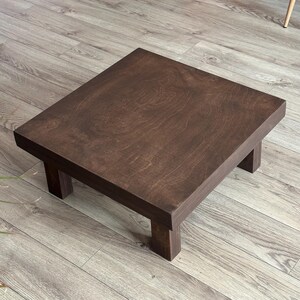 May include: A square, dark brown wooden step stool with four short legs. The stool has a simple, minimalist design and is made of stained wood. The wood grain is visible on the surface.