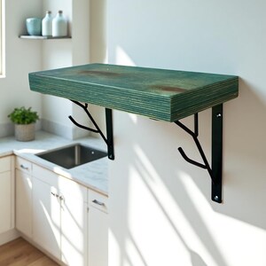 May include: A wall-mounted shelf with a distressed teal finish. The shelf is supported by black metal brackets shaped like tree branches. The shelf is rectangular and appears to be made of wood. The brackets are mounted on a white wall.