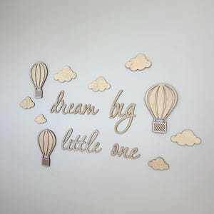 May include: Wooden wall decor featuring hot air balloons, clouds, and the phrase "dream big little one." The natural wood pieces are arranged on a light gray wall, creating a whimsical nursery or children's room theme.