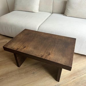 May include: A dark brown wooden coffee table with a rectangular top and angled legs. The table is approximately 76 cm long and 46 cm wide, with a natural wood grain finish. The table is in front of a cream-colored sofa.