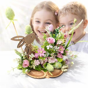 Könnte beinhalten: Ein Osterarrangement mit einem Holzhasen, künstlichen Blumen und dekorativen Eiern. Das Arrangement umfasst rosa Rosen, grünes Laub und ein hellgrünes Ei auf einem Stock. Das Arrangement steht auf einer Holzbasis.