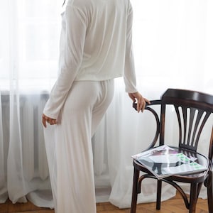 May include: A white, ribbed-knit pajama set. The set includes a long-sleeved top and wide-leg pants. The model is standing near a wooden chair with a magazine on it, next to a window with white curtains.