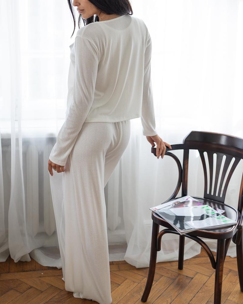 May include: A white, ribbed-knit pajama set. The set includes a long-sleeved top and wide-leg pants. The model is standing near a wooden chair with a magazine on it, next to a window with white curtains.