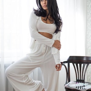 May include: A three-piece white lounge set consisting of a crop top, long-sleeve cardigan, and wide-leg pants. The outfit is made of a soft, ribbed fabric. The model is standing near a window and a wooden chair.