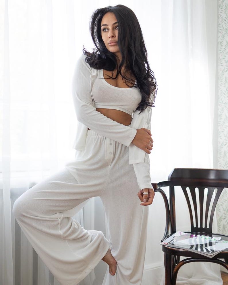 May include: A three-piece white lounge set consisting of a crop top, long-sleeve cardigan, and wide-leg pants. The outfit is made of a soft, ribbed fabric. The model is standing near a window and a wooden chair.