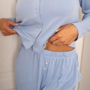 May include: Light blue pajama set. The set includes a long-sleeved button-down shirt and matching pants. The shirt has a ribbed texture and white buttons. The pants have a button closure at the waist. The fabric appears soft and comfortable.
