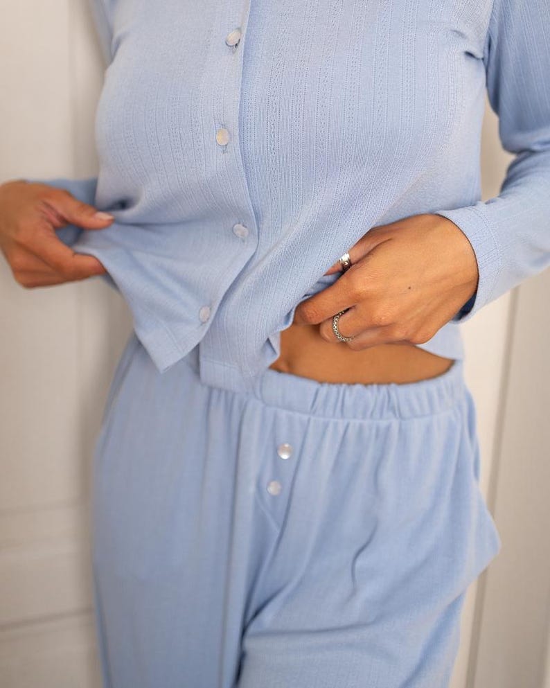 May include: Light blue pajama set. The set includes a long-sleeved button-down shirt and matching pants. The shirt has a ribbed texture and white buttons. The pants have a button closure at the waist. The fabric appears soft and comfortable.