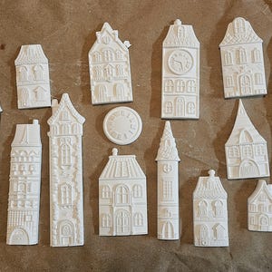 May include: A collection of white, unpainted miniature house and clock tower ornaments. The ornaments vary in size and design, with some featuring intricate architectural details. A ruler is visible on the right side of the image.