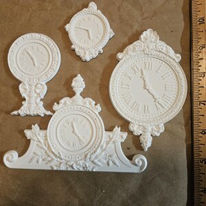 May include: Four white decorative clock molds with Roman numerals and ornate designs. The clocks vary in size and shape, including a round clock on a stand, a square clock, and a large clock with a decorative top. A ruler is in the background.