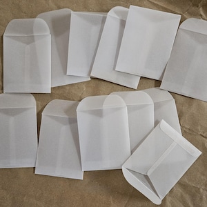 May include: A collection of small, translucent white paper envelopes. The envelopes are rectangular with a curved top flap and are arranged on a brown surface. The envelopes are empty and ready for use.