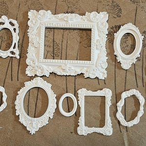 May include: A collection of white, ornate picture frames in various shapes and sizes. The frames include oval, rectangular, and irregular designs, all with detailed, decorative molding. A wooden ruler is visible on the right side.