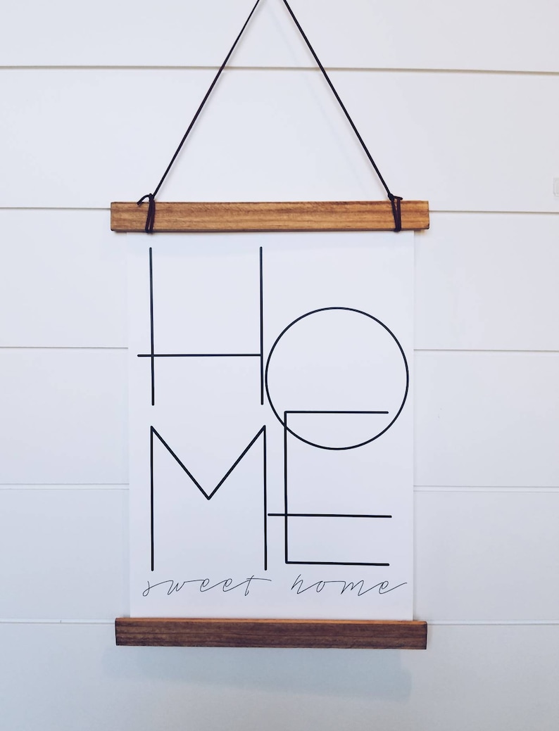 May include: A black and white print with the word "HOME" in a geometric design. The print is framed with a natural wood frame and hanging on a white wall. The text "sweet home" is written below the word "HOME".