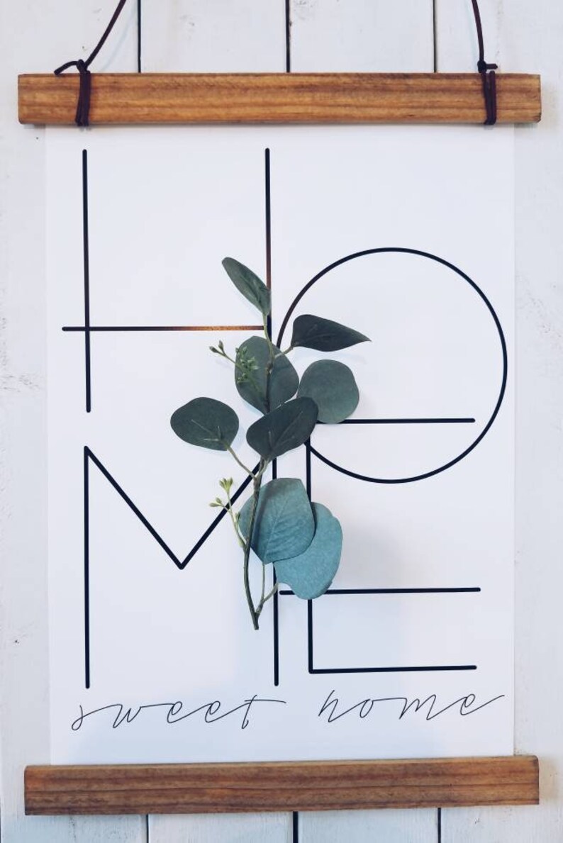 May include: A black and white print with the words "sweet home" written in cursive. The word "home" is formed with geometric shapes and a sprig of green eucalyptus is placed in the center of the print.