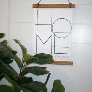 May include: A black and white print with the word "home" written in a geometric style. The print is hanging on a white wall with a wooden frame and brown string.
