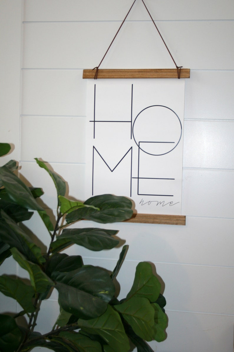 May include: A black and white print with the word "home" written in a geometric style. The print is hanging on a white wall with a wooden frame and brown string.