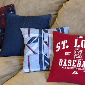 May include: Four decorative pillows on a beige couch. The pillows are made from repurposed shirts. One pillow is red with white text that reads "St. Louis Est. 1892 Baseball MLB Authentic Collection". Another pillow is blue denim with a white button. The third pillow is a blue and white plaid with a white button. The fourth pillow is a red and black plaid with a white button.