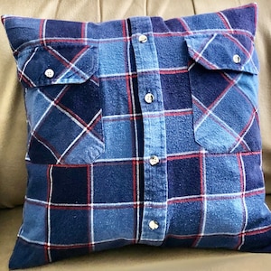 May include: A blue and red plaid pillow with two pockets and buttons down the front. The pillow is made from a repurposed flannel shirt.