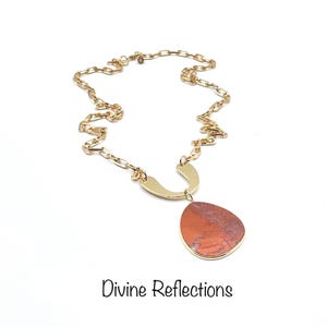 May include: A gold-toned necklace with a large, teardrop-shaped pendant. The pendant is a mix of red and brown tones, set in a gold frame. The chain is a linked design. The text "Divine Reflections" is at the bottom.